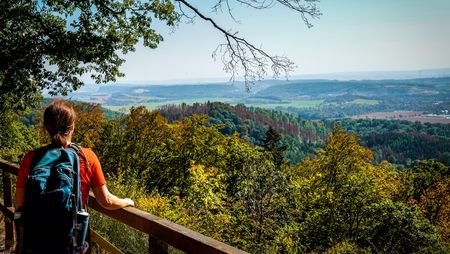 Eine Wandererin mit Rucksack blickt entspannt von einer Holzbrüstung über die herbstliche Südharzer Landschaft. Die weiten, grünen Hügel und der klare Himmel im Hintergrund laden zu Entdeckungen in der idyllischen Natur des Südharzes ein.