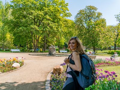 Das Bild zeigt eine Frau mit Rucksack im Vordergrund und einen mit bunten Beeten angelegten Kurpark im Sommer. 