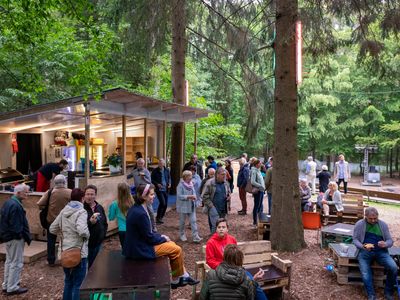 Das Bild zeigt eine Waldbühne mit Menschen und einer Hütte vor grünen Bäumen.