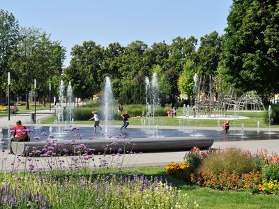 Das Bild zeigt einen Brunnen mit mehreren Fontänen und gesäumt von bunten Blumen im Sommer. 