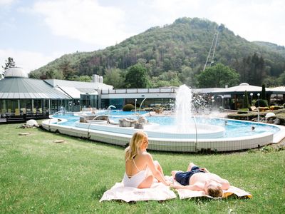 Das Bild zeigt ein Pärchen auf einer grünen Wiese vor einem Außenbecken mit Fontäne der Sole-Therme in Bad Harzburg.