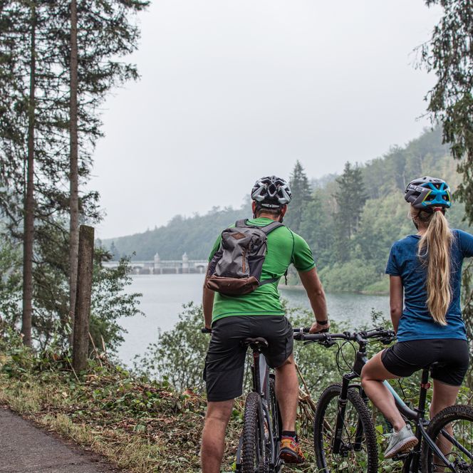 Zwei Radfahrer, eine Frau und ein Mann, stehen auf dem Harzrundweg und blicken auf eine Talsperre.